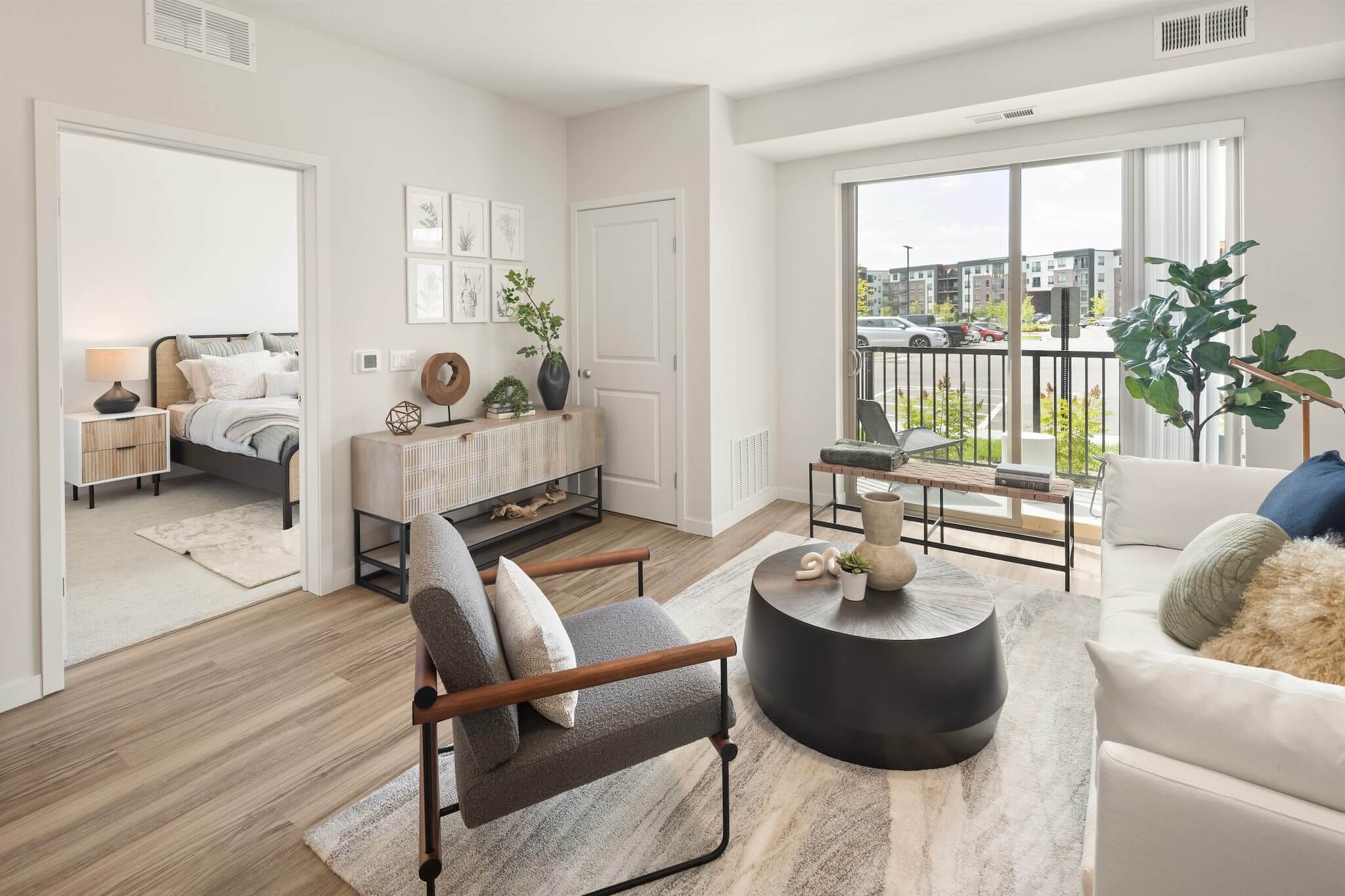 Aster at Riverdale Station apartment living area showing exterior patio door, living room furniture, hardwood flooring, and an entrance to the bedroom